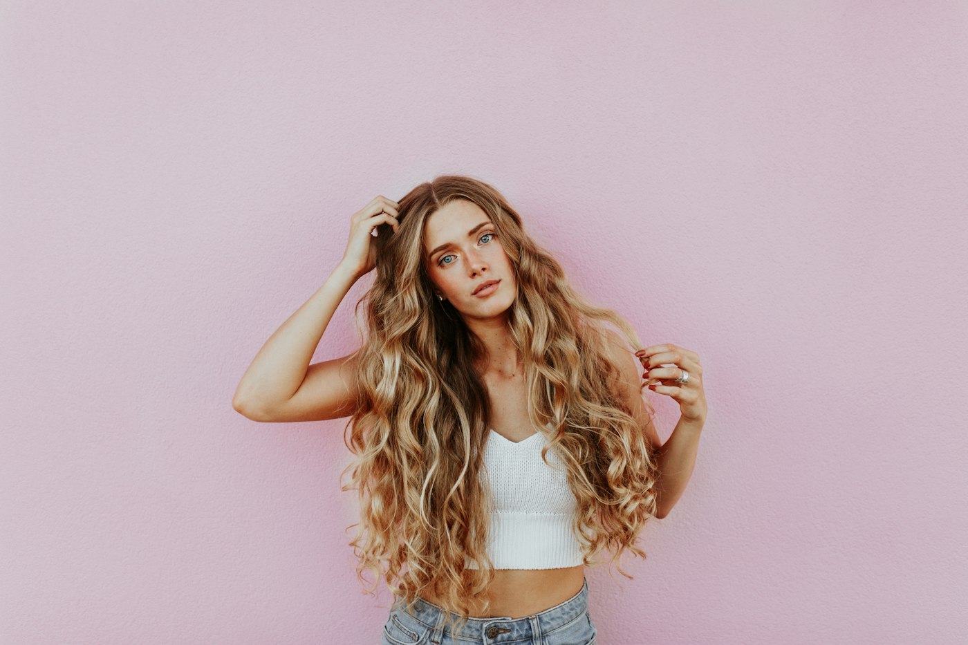 Woman with long wavy hair running her fingers through her hair at the scalp against a pink wall